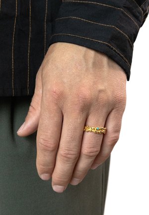 Goldring mit einer strukturierten Band, das zwei kleine blaue Akzente aufweist. Hand ruht auf einem Stoff; sichtbarer Hautton und Nagellackfarbe.
