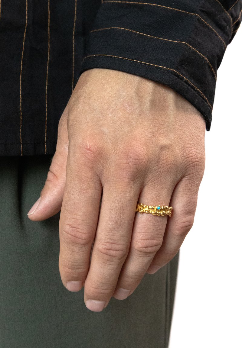Goldring mit einer strukturierten Band, das zwei kleine blaue Akzente aufweist. Hand ruht auf einem Stoff; sichtbarer Hautton und Nagellackfarbe.