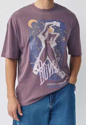 Persoon draagt een paars T-shirt met een abstract dansende figuur, maan, sterren en de tekst "David Bowie Serious Moonlight Tour."
