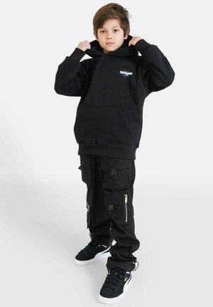Felpa nera con cappuccio e tasca frontale, abbinata a pantaloni cargo neri con più tasche e cerniere. Sneakers nere completano il look.