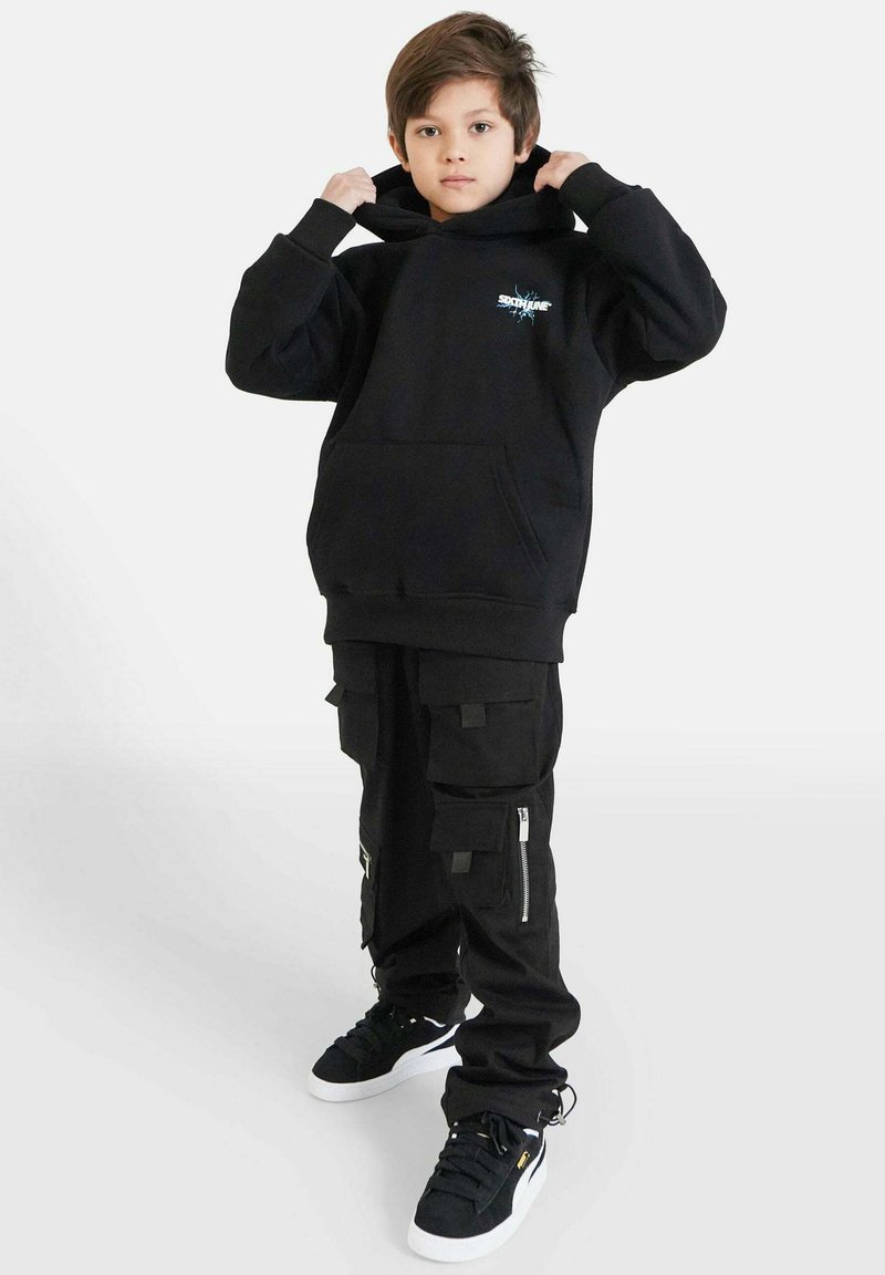 Felpa nera con cappuccio e tasca frontale, abbinata a pantaloni cargo neri con più tasche e cerniere. Sneakers nere completano il look.
