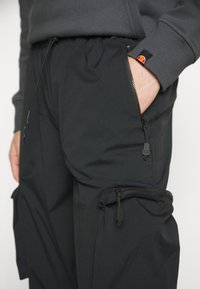 Main dans la poche d'un pantalon cargo noir avec des poches zippées, porté avec un haut à manches longues gris foncé affichant un petit logo orange et rouge.