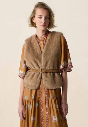 Jonge vrouw met kort blond haar draagt een bruine bontachtige vest met ceintuur in de taille over een gemêleerde mosterdkleurige jurk met halve mouwen.