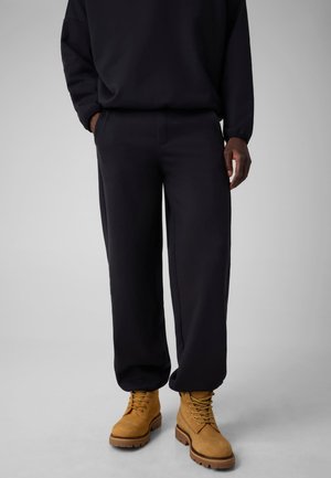 Personne portant un pantalon de survêtement noir et des bottes à lacets jaune moutarde, debout sur une surface grise les mains dans les poches.