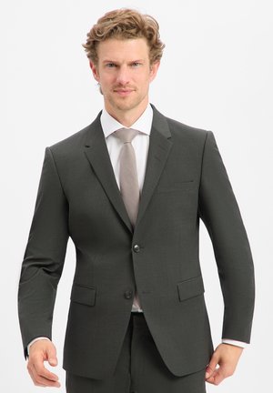 Mann mit hellbraunen Haaren, der einen dunkelgrauen Anzug, ein weißes Hemd und eine hellgraue Krawatte trägt, steht vor weißem Hintergrund.