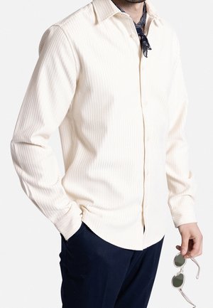 Hombre con camisa cremosa texturizada abotonada, pantalones azul marino, una bufanda oscura con estampado en el cuello, sosteniendo unas gafas de sol con montura plateada en la mano derecha.