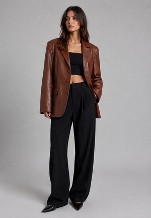 Woman with long dark hair wearing a brown leather blazer, black cropped top, wide black trousers, and black pointed shoes standing against a gray background.
