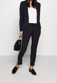 Navyblazer über einem weißen Oberteil, kombiniert mit navyfarbenen Jogginghosen mit weißen Seitenstreifen. Eine schwarze Tasche und Schlupfloafers runden den Look ab.