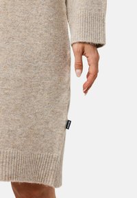 Beige gebreide jurk met een geribbelde zoom, waarin een gestructureerde stof en een label aan de zijkant zijn verwerkt. Close-up van een hand bij de onderkant van het kledingstuk.
