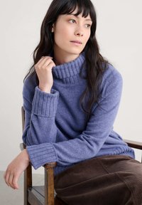 Lila gebreide trui met een hoge ribbelkraag en ribbelmanchetten, gecombineerd met een bruine corduroy rok, zittend op een houten stoel.