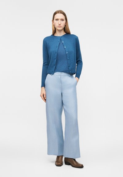 Γυναίκα που φοράει μπλε ζακέτα με κουμπιά πάνω από μπλε τοπ, ανοιχτόχρωμο μπλε παντελόνι με ευρύ πόδι και καφέ μποτάκια μέχρι τον αστράγαλο, στέκεται με το ένα χέρι στην τσέπη.