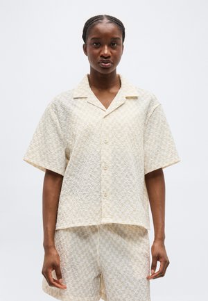 Μαύρη γυναίκα με πλεξούδες, φορώντας κρεμ υφή κοντομάνικο πουκάμισο με κουμπιά και ταιριαστά σορτς, στέκεται μπροστά σε απλό φόντο.