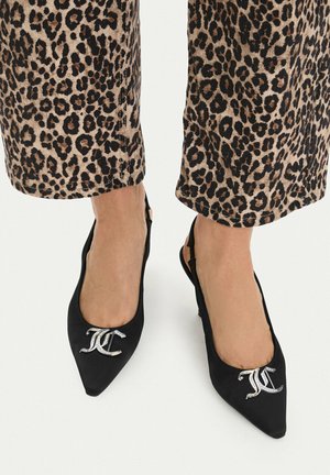 Piedi con scarpe slingback nere a punta con dettaglio logo argento, abbinate a pantaloni corti con stampa leopardata su sfondo bianco.