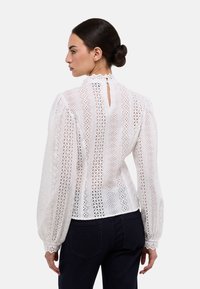 Blouse blanche à manches bouffantes, ornée de détails en dentelle, motifs verticaux, col haut et dos avec ouverture en forme de goutte et fermeture par bouton.