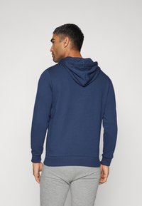 Felpa blu navy con cappuccio regolabile e polsini a costine, abbinata a pantaloni grigi chiari. Il materiale sembra liscio e confortevole.