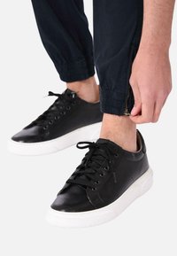 Sneakers in pelle nera con suola in gomma bianca, dotati di chiusura a lacci e perforazioni sui lati. I pantaloni hanno polsini elastici e cerniere.