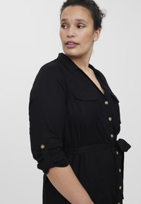 Vestido negro con un frente abotonado, cuello de solapa, dos bolsillos en el pecho, mangas enrolladas y cintura atada. Textura de tejido suave, diseño minimalista.