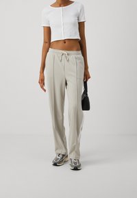 Bílý zkrácený button-up top, béžové kalhoty s elastickým pasem a bočními pruhy, a vzorované sportovní boty; nese malou černou kabelku.