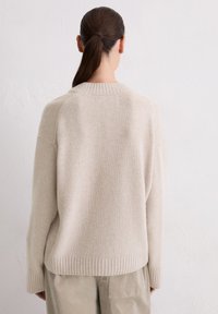 Beige gebreide trui met een ontspannen pasvorm, geribde kraag en zoom, verlaagde schouders en een zachte textuur, gecombineerd met lichtbeige broek.