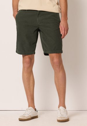 Mann trägt dunkelgrüne knielange Shorts und weiße Sneaker und steht vor einem schlichten Hintergrund.