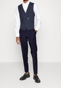Marine vest met dubbele knopen, zilveren ritsdetail, gecombineerd met slim-fit marineblauwe broek en zwarte leren instappers. Witte shirt eronder.