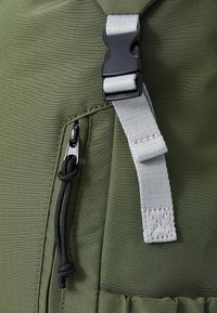 Gros plan sur un tissu vert olive avec une tirette de fermeture éclair noire et une sangle grise ajustable avec une boucle en plastique noir.