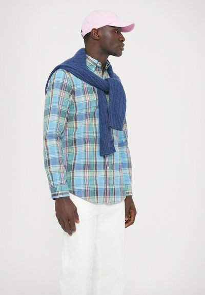 Karirana srajca v modrih, zelenih in rožnatih odtenkih, kombinirana z modrim pletenim puloverjem, ki je premeten čez ramena, in rožnato kapo. Bele hlače.