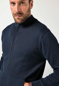 Granatowy sweter na suwak z prążkowanym wysokim kołnierzem, fakturowanymi panelami na piersi i bocznymi kieszeniami. Miękki, lekki materiał o gładkim wykończeniu.