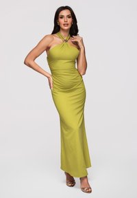 Femme aux cheveux foncés portant une longue robe dos nu vert citronnier, avec des boucles d'oreilles dorées et des talons ouverts, posant avec une main sur la hanche.