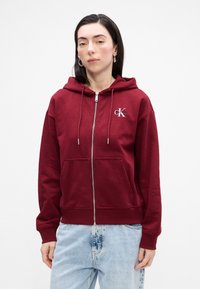 Bordo dukserica s patentnim zatvaračem od mekane tkanine, s prednjim džepom, kapuljačom s vezicom i logom na lijevoj strani prsnog koša. Uparena s svijetlim trapericama.