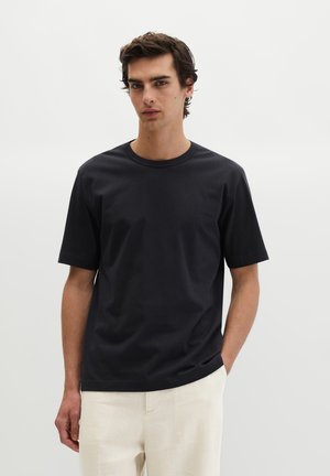 Jeune homme aux cheveux foncés portant un t-shirt noir uni à manches courtes et un pantalon beige clair, debout sur un fond blanc.