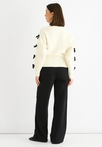 Cream knit sweater featuring black ribbon accents along the sleeves, a fitted ribbed waistband, and a relaxed fit completed with black wide-leg pants.