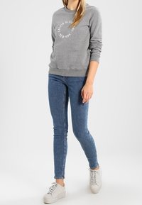 Felpa grigia con scollatura rotonda e stampa del logo bianca, abbinata a jeans blu a sigaretta e sneakers bianche. Tessuto dalla consistenza morbida.