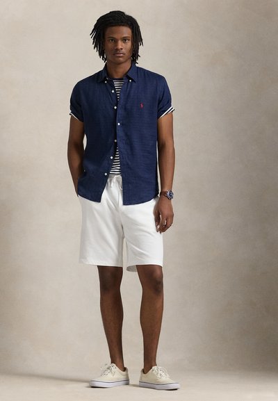 Marineblå kortermet skjorte med knapper og rød logo, stripete undertrøye, hvite shorts og beige sneakers. Glatt stoff, avslappet passform.