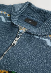 Kék kötött, cipzáros kardigán magas gallérral, ezüst cipzárral és narancssárga, valamint világoskék mintázattal. Címke látható.