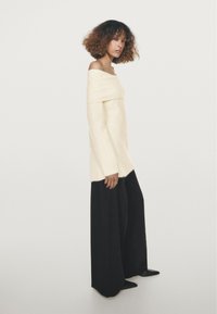 Pull crème clair à épaules dénudées avec une texture douce, associé à un pantalon large noir taille haute et des chaussures noires à bouts pointus.