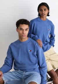 Sudaderas de algodón de color azul claro con mangas largas, con un pequeño logo en el pecho. El modelo masculino lleva pantalones cortos de mezclilla, mientras que la modelo femenina viste una falda beige.