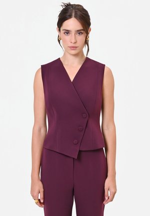 Donna che indossa un gilet su misura senza maniche color prugna con tre bottoni e pantaloni coordinati a vita alta su sfondo neutro.