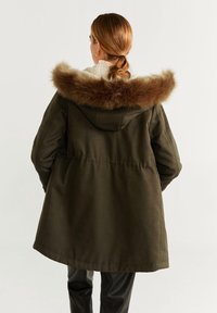 Oliwkowa parka z kapturem wyściełanym futrem, z taliowanym pasem i długimi rękawami. Materiał wydaje się gładki, o matowym wykończeniu.