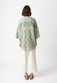 Top verde largo con motivo, dotato di maniche larghe e decorazioni intricate in vari colori. Indossato con pantaloni cropped color crema e sandali.