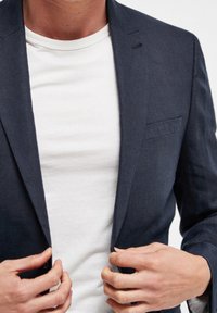 Granatowa lniana marynarka z klapami typu notch, jedną kieszenią na piersi i dopasowanym krojem, noszona na białej koszulce z okrągłym dekoltem.