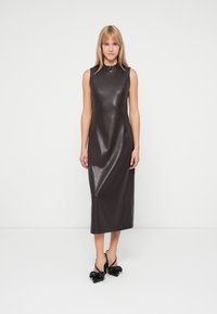 Vrouw staat in een mouwloze, donkerbruine, glanzende midi-jurk met hoge hals en zwarte, puntige hakken met strikken.