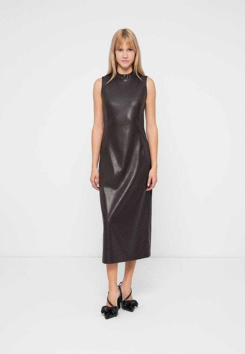 Vrouw staat in een mouwloze, donkerbruine, glanzende midi-jurk met hoge hals en zwarte, puntige hakken met strikken.