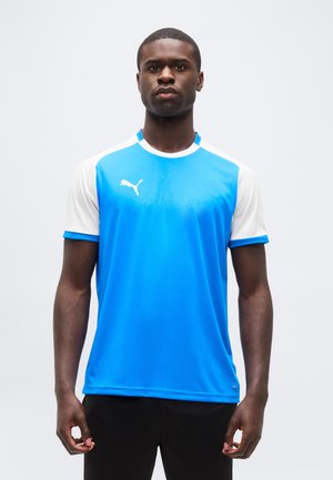 Fiatal férfi áll, kék-fehér Puma sportinget és fekete nadrágot visel, egyenes előre néz egy egyszerű háttér előtt.