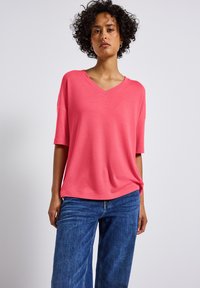 Růžový top s V-výstřihem a krátkým rukávem, s volným střihem, vyrobený z měkké látky. V kombinaci s modrými džínovými kalhotami širokého střihu. Jednoduchý a moderní design.