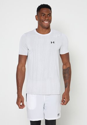 Mężczyzna uśmiechający się, ubrany w białą koszulkę Under Armour z krótkim rękawem, białe spodenki oraz czarne spodenki pod spodem, pokazujący tatuaż wilka na lewym przedramieniu.