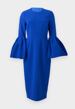 Nő áll, kezét a csípőjén tartva, elektromos kék midi ruhában, amelynek hosszú, bővülő ujjai vannak, fekete magas sarkú szandálban, fehér háttér előtt.