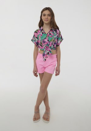 Camicetta nera a maniche corte con bottoni, decorata da un colorato motivo a foglie tropicali, abbinata a pantaloni corti rosa chiaro e sandali argentati.