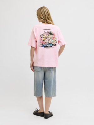 Osoba z długimi blond włosami ubrana w różowy t-shirt z kolorowym nadrukiem Ibiza i jasnoniebieskie szorty jeansowe, stojąca na białym tle.