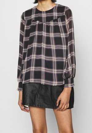 Kobieta ubraną w czarno-beżową kratowaną bluzkę z długim rękawem, połączoną z czarną skórzaną mini spódnicą, stoi na jednolitym tle.